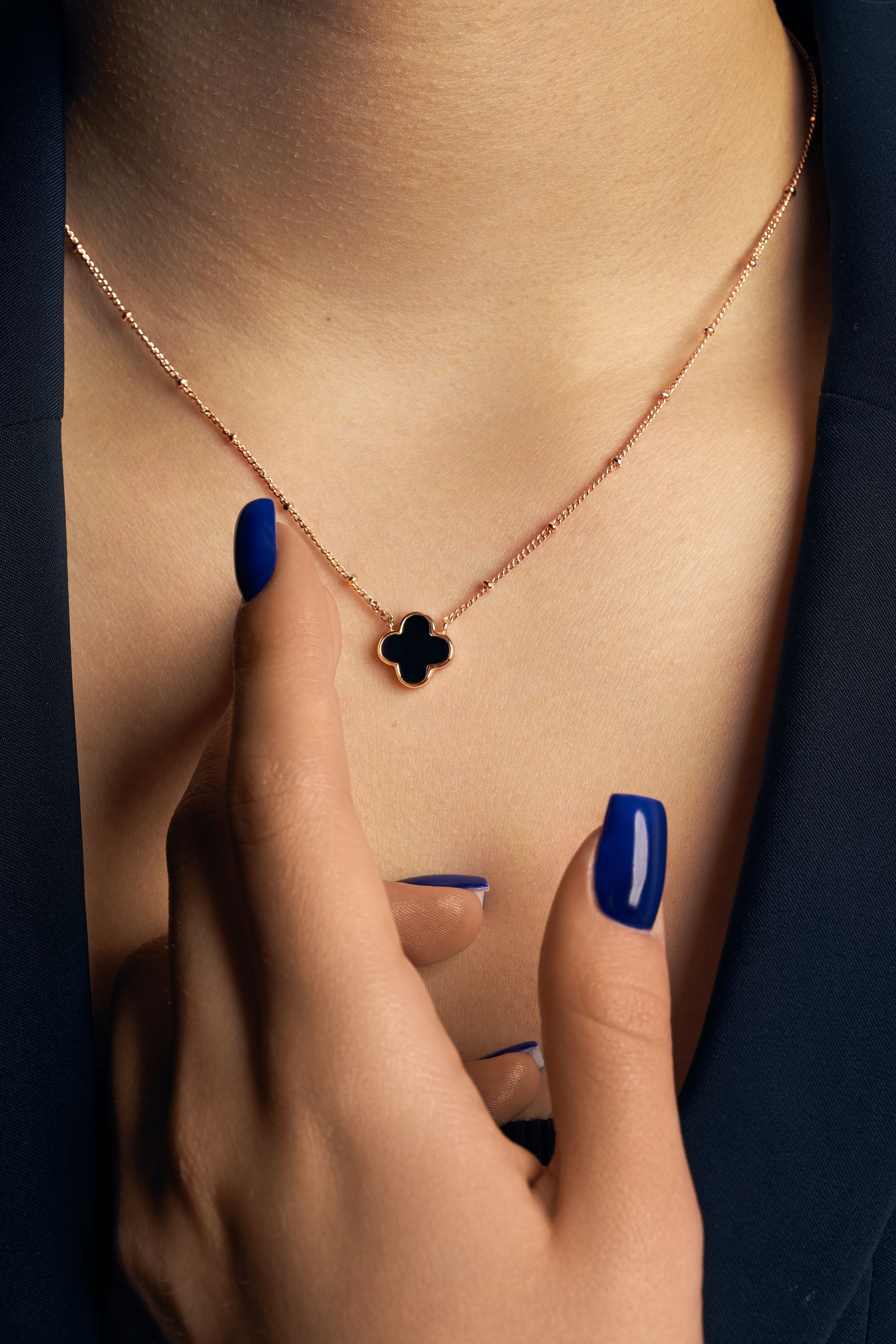 A close-up shot of a woman's neck and hand, featuring a delicate gold necklace with a black clover-shaped pendant. van kleef and arpel affordable luxury jewellery womens mens kings queens white gold silver gold diamond gemstone precious stone chains and necklaces earrings watches rings exclusive by Deep Shine Jewellery jewellery jewelry DSJ Your Time To Shine